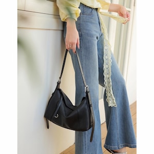 May include: A black leather shoulder bag with a curved design. The bag has a long, adjustable strap and a silver-tone buckle. The bag is held by a person wearing blue jeans and a yellow top.