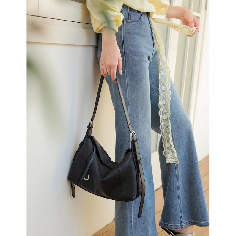 May include: A black leather shoulder bag with a curved design. The bag has a long, adjustable strap and a silver-tone buckle. The bag is held by a person wearing blue jeans and a yellow top.
