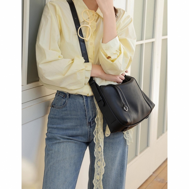 May include: A black leather shoulder bag with a silver-tone ring detail. The bag is carried by a person wearing a light yellow button-down shirt and blue jeans. The outfit is accessorized with a black strap and a pair of light yellow glasses.