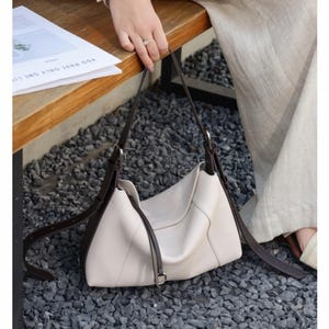 May include: Cream-colored shoulder bag with dark brown straps and accents. The bag has a curved shape and a minimalist design. The straps are long and adjustable. The bag is sitting on a bed of gray gravel.
