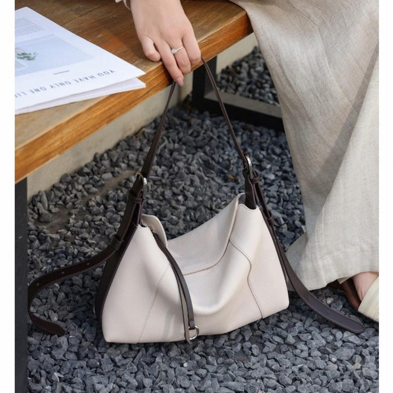 May include: Cream-colored shoulder bag with dark brown straps and accents. The bag has a curved shape and a minimalist design. The straps are long and adjustable. The bag is sitting on a bed of gray gravel.