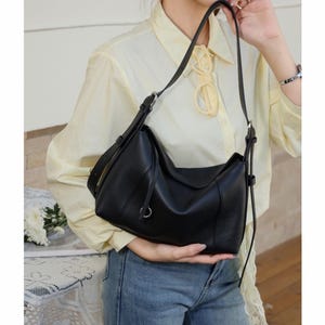 May include: Black leather shoulder bag with a curved design. The bag has a long, adjustable strap and a small silver-tone ring detail. The bag is held by a person wearing a yellow shirt and blue jeans.