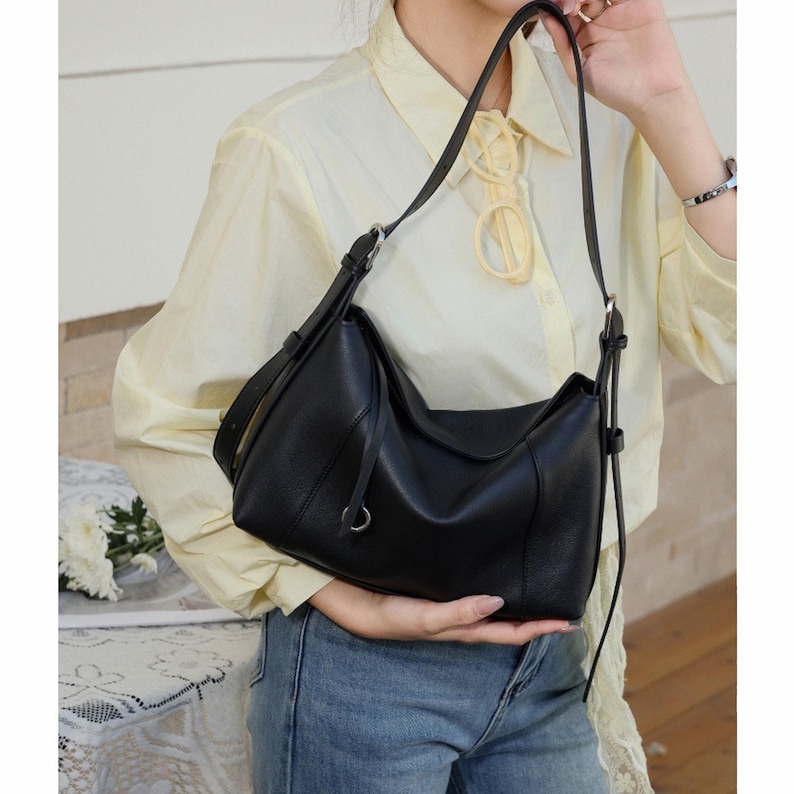 May include: Black leather shoulder bag with a curved design. The bag has a long, adjustable strap and a small silver-tone ring detail. The bag is held by a person wearing a yellow shirt and blue jeans.