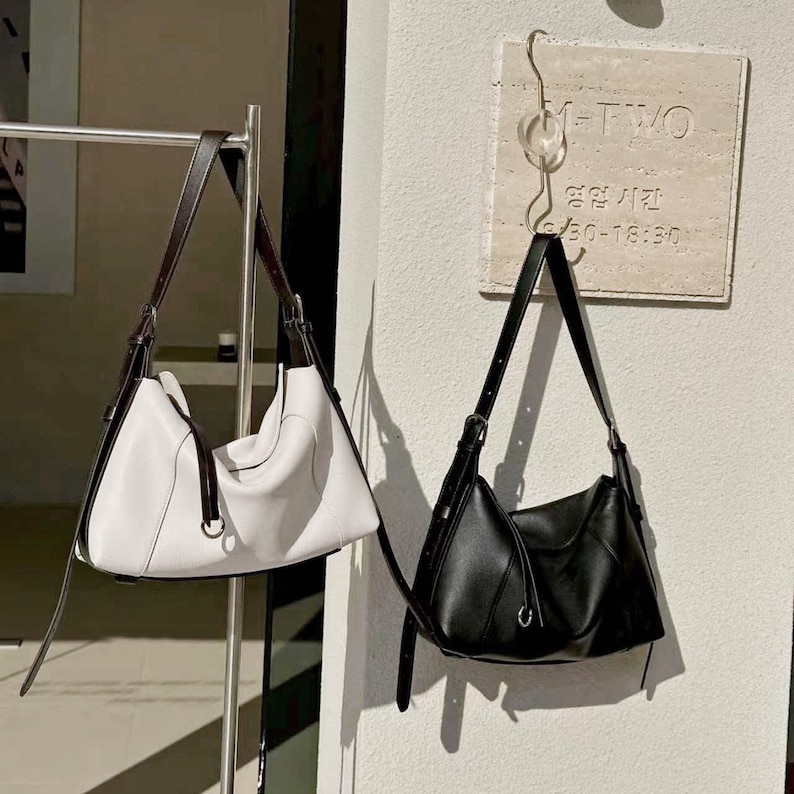 May include: Two leather shoulder bags, one white and one black, hang against a white wall. The white bag has brown straps and a silver ring detail. The black bag has black straps and a silver ring detail. The bags are similar in shape and size.