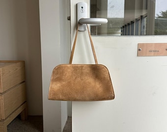 May include: A tan suede handbag with a structured, semi-circular silhouette and two thin, matching shoulder straps. The bag features a soft, textured surface and a clean, minimalist design with visible stitching along the edges of the main compartment.