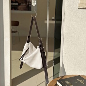 May include: A white leather shoulder bag with dark brown straps and silver-tone hardware is suspended from a metal hook. The bag has a soft, slouchy design and a minimalist aesthetic. The straps are adjustable and appear to be made of a smooth leather material.