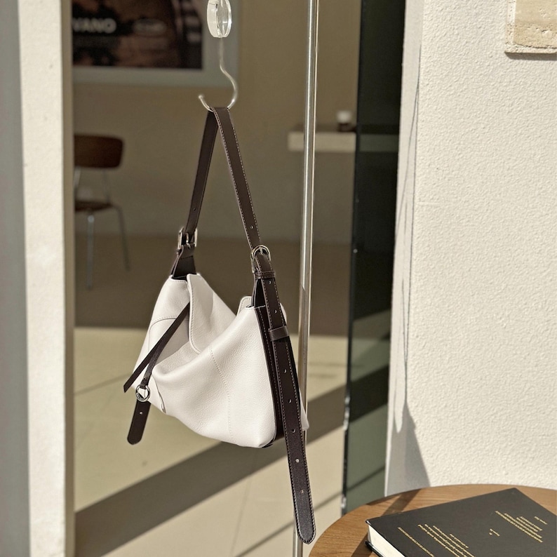 May include: A white leather shoulder bag with dark brown straps and silver-tone hardware is suspended from a metal hook. The bag has a soft, slouchy design and a minimalist aesthetic. The straps are adjustable and appear to be made of a smooth leather material.