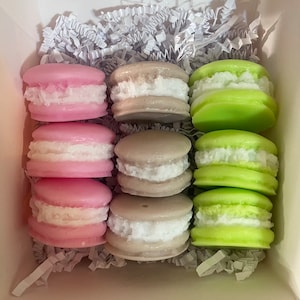 May include: A box containing nine macaron-shaped candles. The candles are in three colours: pink, green, and grey. Each candle has a white filling.