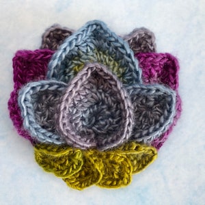 May include: A handmade crochet flower in shades of purple, blue, gray, and green. The flower has multiple layers of petals, with a textured appearance. The petals are arranged in a circular pattern, creating a three-dimensional effect.