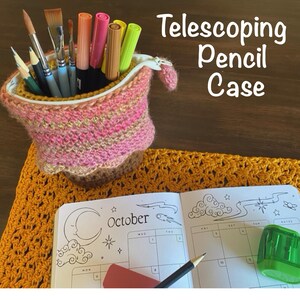 May include: A crocheted telescoping pencil case in pink, brown, and gold, filled with art supplies. The case sits on a yellow crocheted placemat next to an open October calendar with a pencil, eraser, and sharpener. The text "Telescoping Pencil Case" is visible.