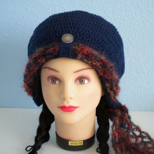 Op de afbeelding: Een blauwe gebreide beanie met een kleurrijke gebreide rand en een zilveren knoop. De beanie zit op een mannequinhoofd met zwarte vlechten.