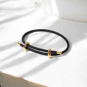 Könnte beinhalten: Ein schwarzes geflochtenes Armband aus Seil mit goldenen Metallakzenten. Das Armband hat einen verschiebbaren Knotenverschluss.