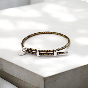 Könnte beinhalten: Ein geflochtenes Armband in Schwarz und Gelb mit silbernen Akzenten. Das Armband hat einen runden silbernen Anhänger und drei silberne Ringe.