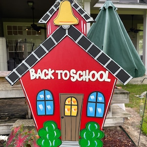 May include: A red wooden schoolhouse sign featuring a yellow bell at the top, black roof shingles, two blue windows, a brown door, and green bushes at the base. The text BACK TO SCHOOL is written in white letters across the center of the sign.