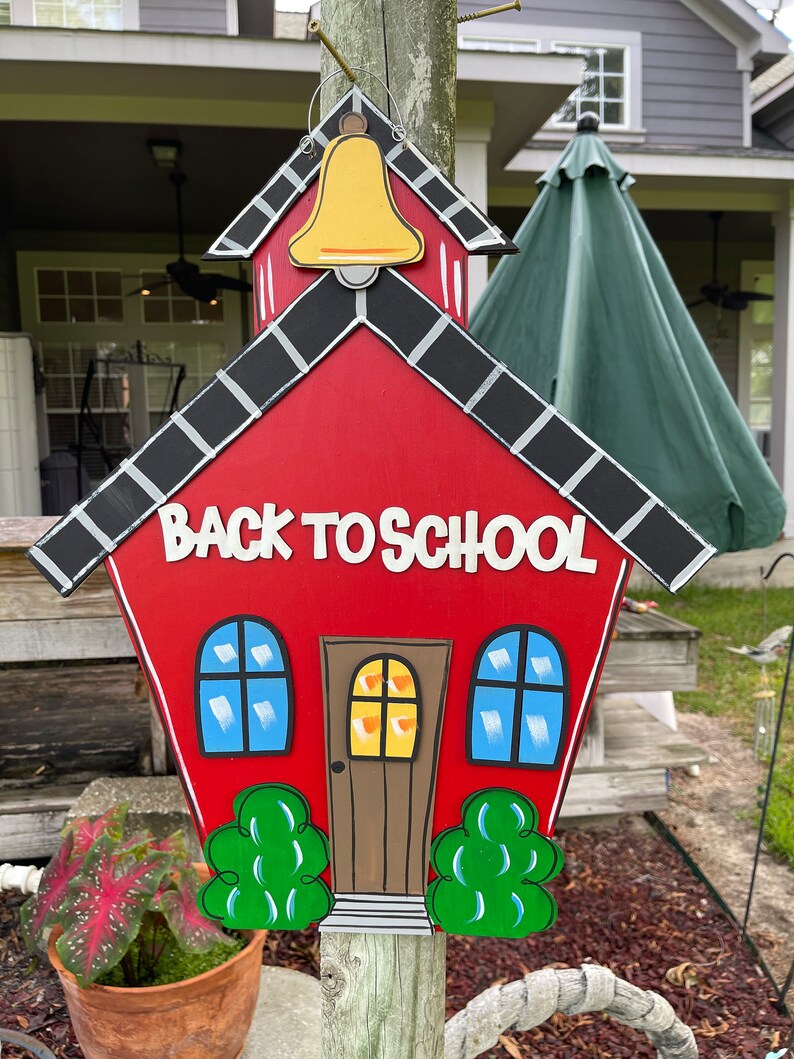 May include: A red wooden schoolhouse sign featuring a yellow bell at the top, black roof shingles, two blue windows, a brown door, and green bushes at the base. The text BACK TO SCHOOL is written in white letters across the center of the sign.