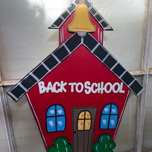 May include: A red wooden schoolhouse wall hanging featuring a gold bell at the top, black and white roof shingles, and the text BACK TO SCHOOL in white letters. The design includes a brown door, two blue windows, and green bushes at the base.