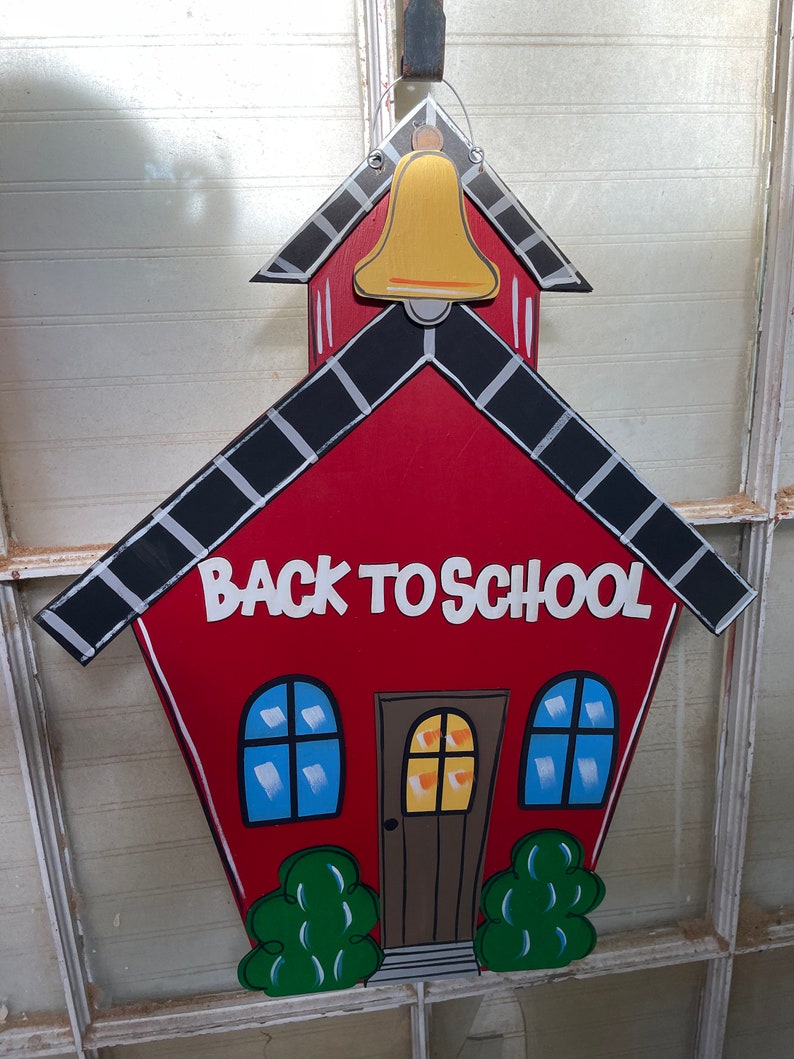 May include: A red wooden schoolhouse wall hanging featuring a gold bell at the top, black and white roof shingles, and the text BACK TO SCHOOL in white letters. The design includes a brown door, two blue windows, and green bushes at the base.