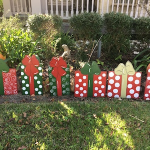 May include: Wooden Christmas present cutouts in a row, painted red, green, and white. Each gift has a large bow and decorative patterns, including polka dots and swirls. The gifts are displayed outdoors in a grassy area.