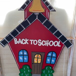 May include: A red schoolhouse-shaped wooden sign featuring a yellow bell at the top, black roof trim, and the white text BACK TO SCHOOL. The design includes a brown door, two blue windows, and two green bushes at the base, all with black outlines.