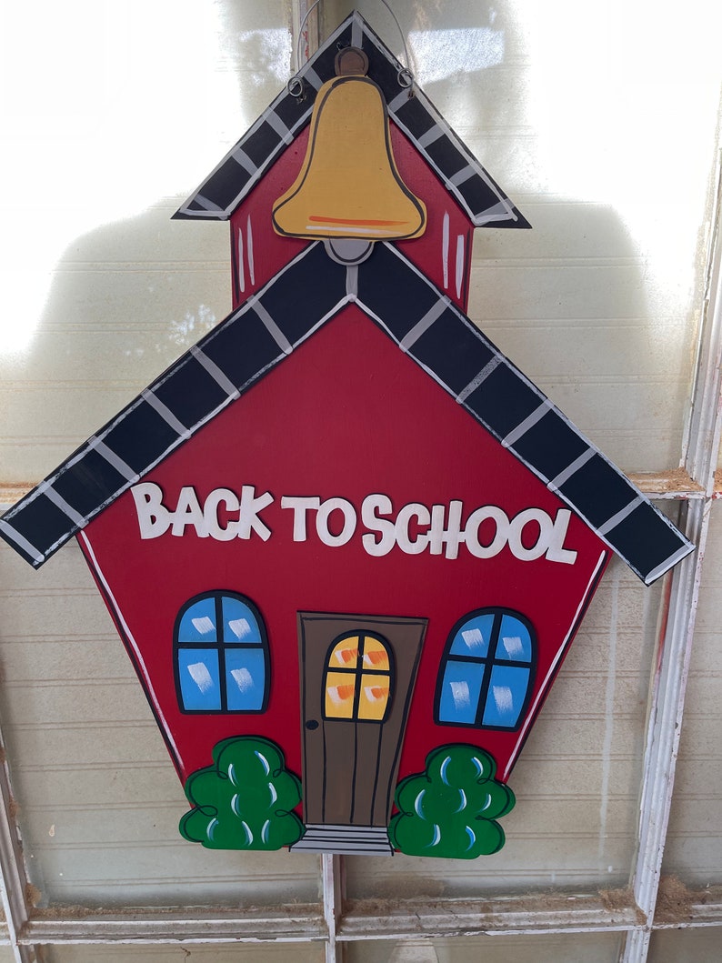 May include: A red schoolhouse-shaped wooden sign featuring a yellow bell at the top, black roof trim, and the white text BACK TO SCHOOL. The design includes a brown door, two blue windows, and two green bushes at the base, all with black outlines.