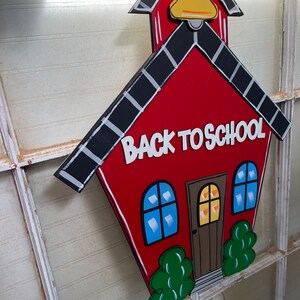 May include: A red schoolhouse-shaped wooden sign featuring a yellow bell at the top, black roof shingles with white accents, two blue windows, a brown door, and green bushes. The text BACK TO SCHOOL is printed in white letters on the front.