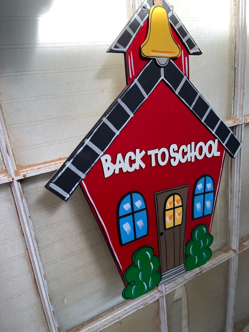 May include: A red schoolhouse-shaped wooden sign featuring a yellow bell at the top, black roof shingles with white accents, two blue windows, a brown door, and green bushes. The text BACK TO SCHOOL is printed in white letters on the front.