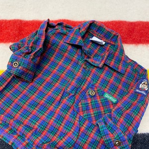 Puede incluir: Camisa de franela a cuadros rojos, azules y verdes con mangas largas y cierre de botones. La camisa tiene un parche en el pecho izquierdo que dice "OshKosh" y otro parche en el pecho derecho que dice "Junior Mountain" con un oso que lleva un sombrero de guardabosques.
