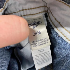 Puede incluir: Primer plano de una etiqueta blanca de ropa Oshkosh B'gosh. La etiqueta indica la talla 24M e incluye el texto "MADE IN BANGLADESH". Tambi&eacute;n se proporcionan instrucciones de lavado. La etiqueta est&aacute; adherida a material vaquero.