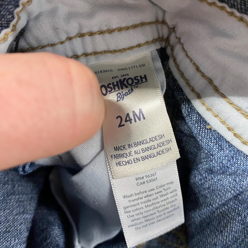 Puede incluir: Primer plano de una etiqueta blanca de ropa Oshkosh B'gosh. La etiqueta indica la talla 24M e incluye el texto "MADE IN BANGLADESH". Tambi&eacute;n se proporcionan instrucciones de lavado. La etiqueta est&aacute; adherida a material vaquero.