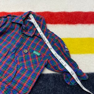 Puede incluir: Camisa de franela a cuadros rojos, azules y verdes de manga larga y cierre de botones. La camisa tiene un bolsillo en el pecho izquierdo con el logotipo de OshKosh B'Gosh. La camisa tiene un parche en la manga derecha con las palabras "Hecho en China".