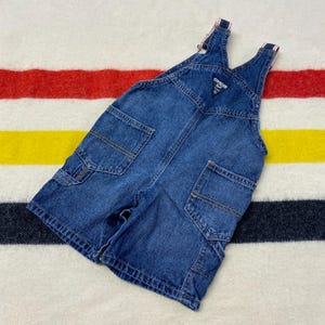 Puede incluir: Peto corto vaquero azul OshKosh B'gosh. El peto tiene m&uacute;ltiples bolsillos, un bolsillo en el peto y tirantes ajustables con detalles a rayas rojas y blancas. La marca es visible en el peto. La prenda est&aacute; sobre una manta a rayas.