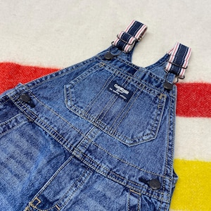 Puede incluir: Primer plano de un peto vaquero azul con tirantes ajustables con rayas rojas y blancas. El peto tiene un bolsillo delantero y la marca "OshKosh B'gosh" es visible en una etiqueta. La prenda est&aacute; sobre un fondo a rayas.