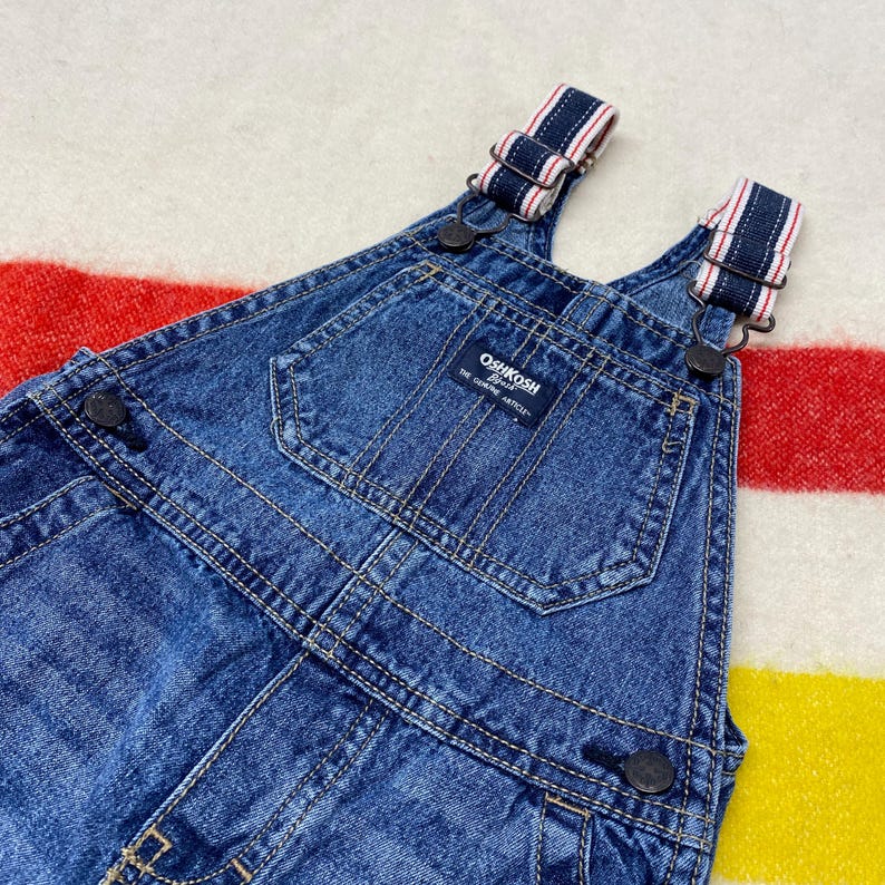 Puede incluir: Primer plano de un peto vaquero azul con tirantes ajustables con rayas rojas y blancas. El peto tiene un bolsillo delantero y la marca "OshKosh B'gosh" es visible en una etiqueta. La prenda est&aacute; sobre un fondo a rayas.