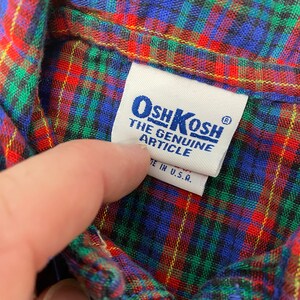 Puede incluir: Una etiqueta OshKosh azul y blanca con el texto "The Genuine Article" y "Made in U.S.A.". La etiqueta est&aacute; unida a una tela de cuadros rojos, verdes y azules.