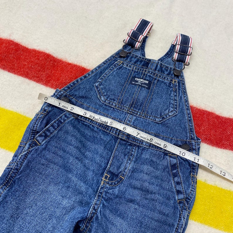 Puede incluir: Peto Oshkosh de mezclilla azul con tirantes a rayas. Una cinta m&eacute;trica muestra que el peto mide aproximadamente 33 cm de largo. El peto tiene un bolsillo delantero y el logotipo de Oshkosh.