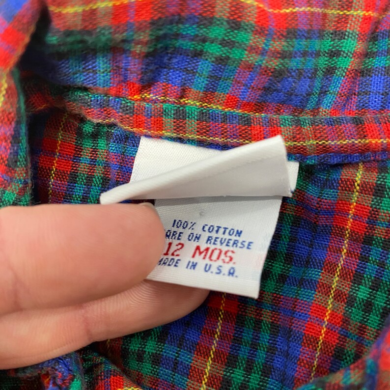 Puede incluir: Una etiqueta blanca con texto rojo y azul que dice "100% algod&oacute;n cuidado en el reverso 12 MOS. hecho en EE. UU." La etiqueta est&aacute; unida a una tela de cuadros rojos, verdes, azules y amarillos.