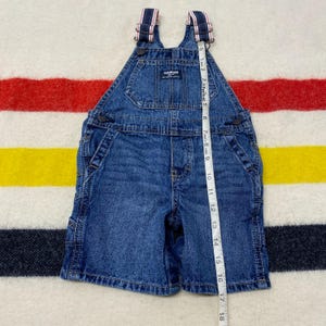 Puede incluir: Mono vaquero azul con tirantes a rayas. El mono tiene un bolsillo de peto, bolsillos laterales y cierre de bot&oacute;n. El mono mide aproximadamente 43 cm de largo.