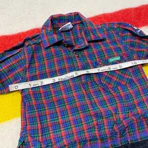 Puede incluir: Camisa de botones a cuadros rojos, azules y verdes con un bolsillo en el lado izquierdo. La camisa tiene una etiqueta que dice "OSK KOSH" y "MADE IN CHINA".