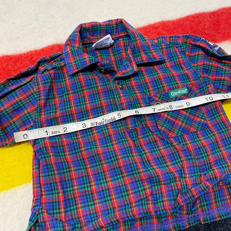 Puede incluir: Camisa de botones a cuadros rojos, azules y verdes con un bolsillo en el lado izquierdo. La camisa tiene una etiqueta que dice "OSK KOSH" y "MADE IN CHINA".