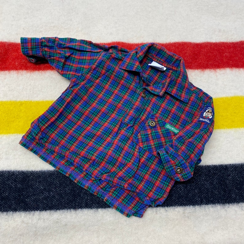 Puede incluir: Camisa de manga larga a cuadros rojos, azules y verdes con cuello abotonado y un bolsillo de parche en el lado izquierdo. La camisa tiene un peque&ntilde;o logotipo de oso bordado en la manga derecha que dice "Oz Koz" y "Seattle".