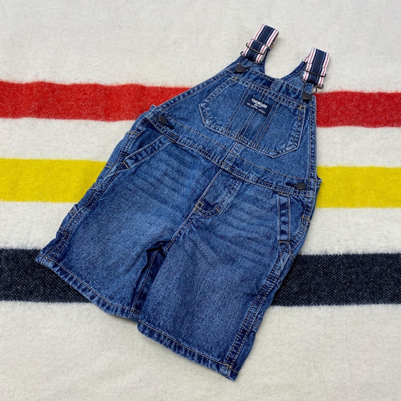 Puede incluir: Pantalones cortos con peto de mezclilla azul con tirantes ajustables con rayas rojas y blancas. Los pantalones cortos tienen m&uacute;ltiples bolsillos y cierres de bot&oacute;n met&aacute;licos. La prenda est&aacute; hecha de mezclilla duradera.