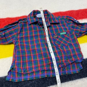 Puede incluir: Camisa de manga larga a cuadros rojos, azules y verdes con un bolsillo en el lado izquierdo. La camisa tiene un cuello abotonado y una etiqueta que dice "OshKosh B'Gosh" en el bolsillo. La camisa se mide con una cinta m&eacute;trica.