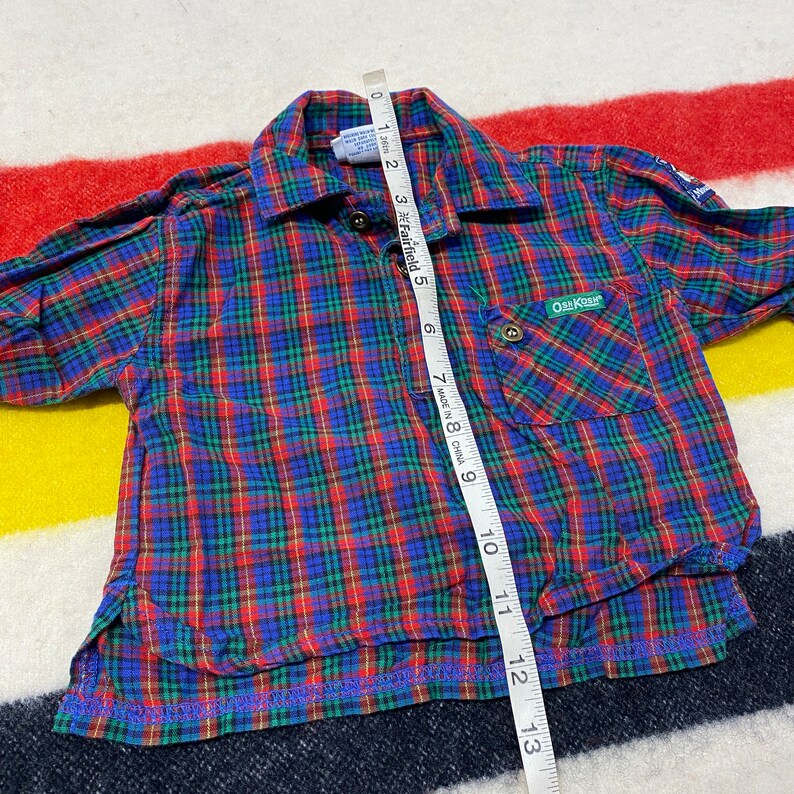 Puede incluir: Camisa de manga larga a cuadros rojos, azules y verdes con un bolsillo en el lado izquierdo. La camisa tiene un cuello abotonado y una etiqueta que dice "OshKosh B'Gosh" en el bolsillo. La camisa se mide con una cinta m&eacute;trica.