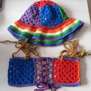 Op de afbeelding: Een regenbooggebreide hoed en bijpassend topje. De hoed is een brede zonnehoed met een gehaakt ontwerp in tinten rood, oranje, geel, groen, blauw en paars. Het topje is halter-stijl met een gehaakt ontwerp in dezelfde kleuren.