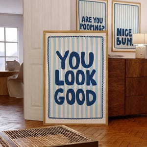 Pode incluir: Três impressões de arte emolduradas com frases humorísticas. A impressão maior diz "YOU LOOK GOOD" em letras azul marinho sobre um fundo listrado azul claro e branco. Duas impressões menores dizem "ARE YOU POOPING?" e "NICE BUM."