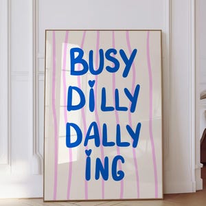 May include: A framed art print with the words "BUSY DILLY DALLY ING" in blue, set against a cream background with vertical pink stripes. The artwork is displayed in a room with a wooden floor.