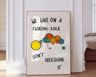 May include: A white poster with a colorful illustration of planets and a cartoon character holding a camera. The text on the poster reads "We live on a floating rock don't overthink it."