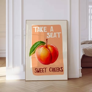 Puede incluir: Impresión artística enmarcada con una vibrante ilustración de melocotón y una hoja verde. El texto "TAKE A SEAT" está encima del melocotón y "SWEET CHEEKS" debajo, sobre un fondo color melocotón. Marco dorado.