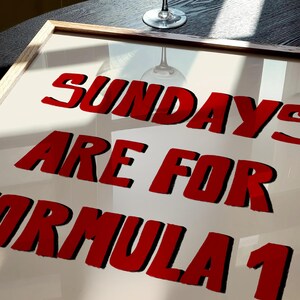 Op de afbeelding: Een ingelijste print met de tekst "SUNDAYS ARE FOR FORMULA 1" in rode, vette letters met zwarte contouren. Een wijnglas is zichtbaar op een donkere houten tafel op de achtergrond.