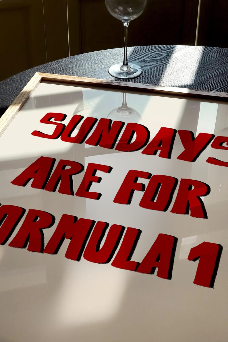 Op de afbeelding: Een ingelijste print met de tekst "SUNDAYS ARE FOR FORMULA 1" in rode, vette letters met zwarte contouren. Een wijnglas is zichtbaar op een donkere houten tafel op de achtergrond.